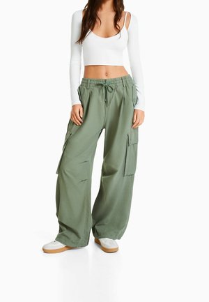Mujer que lleva pantalones cargo holgados color verde oliva, una blusa blanca de manga larga recortada con una tira y zapatillas blancas con suelas de goma.