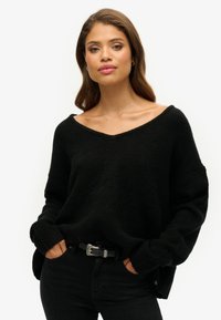 Czarny, oversize sweter z dekoltem w serek, długimi rękawami i prążkowanymi mankietami, zestawiony z czarnymi dżinsami i paskiem z srebrnymi akcentami. Teksturowany materiał.