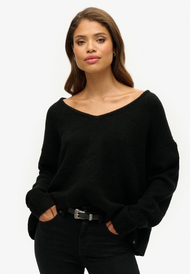 Czarny, oversize sweter z dekoltem w serek, długimi rękawami i prążkowanymi mankietami, zestawiony z czarnymi dżinsami i paskiem z srebrnymi akcentami. Teksturowany materiał.