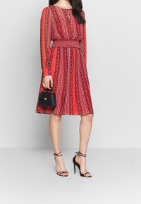 Femme en robe midi à manches longues à motifs rouges, tenant un petit sac à main noir et portant des sandales à talons hauts avec lanières noires, se tenant sur un fond uni.