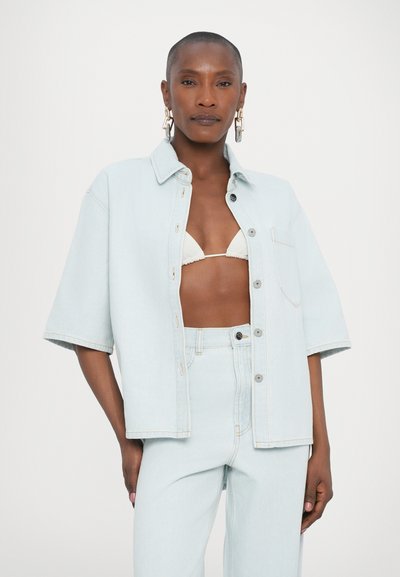 Camisa de ganga azul clara com mangas curtas, botões frontais e um único bolso no peito, combinada com jeans azul claros correspondentes.