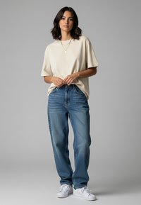 Beige Baumwoll-T-Shirt kombiniert mit blauen Jeans. Das T-Shirt hat einen lockeren Schnitt; die Jeans sind im Straight-Leg-Stil mit einem klassischen Fünf-Taschen-Design. Weiße Sneakers.