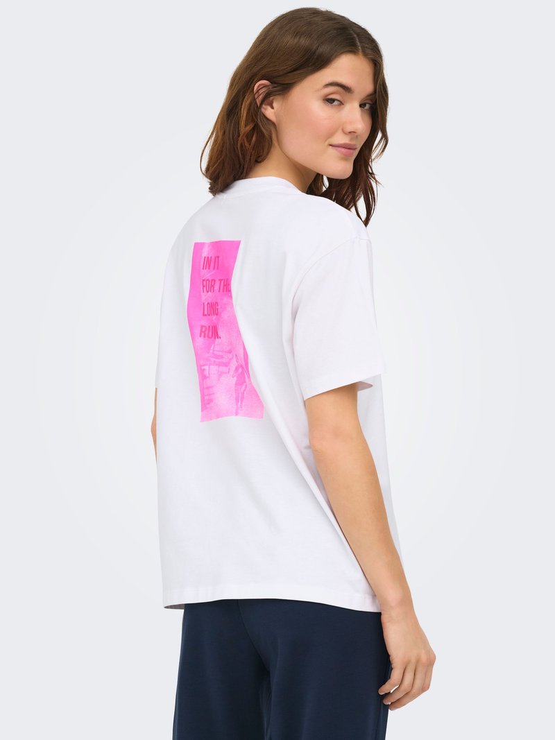 Femme aux cheveux bruns portant un t-shirt blanc surdimensionné avec un texte rectangulaire rose au dos et un pantalon foncé, regardant par-dessus son épaule.