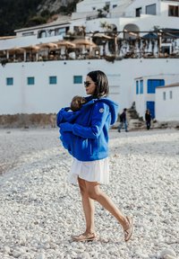Eine blaue Fleece-Hoodie, getragen von einer Frau, mit einer Fronttasche, kombiniert mit einem weißen Kleid und flachen Schuhen, die an einem Kieselstrand spaziert.