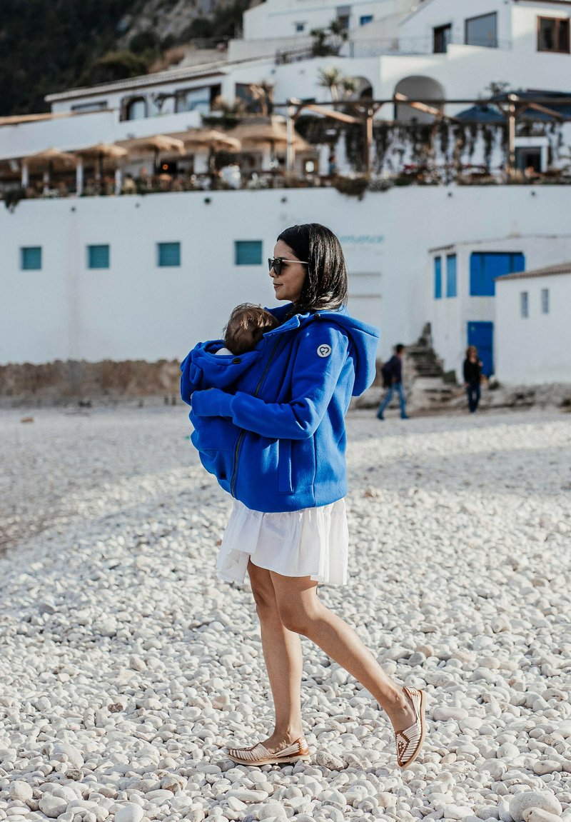 Eine blaue Fleece-Hoodie, getragen von einer Frau, mit einer Fronttasche, kombiniert mit einem weißen Kleid und flachen Schuhen, die an einem Kieselstrand spaziert.
