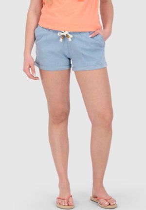 Frau trägt hellblaue Shorts mit umgeschlagenem Saum und einem Seilgürtel, ein oranges Oberteil und korallenfarbene Flip-Flops, steht mit einer Hand in der Tasche.