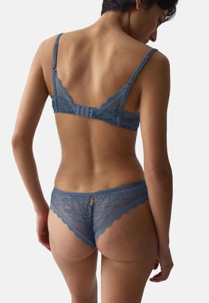 Mujer vista desde atrás con un conjunto de lencería azul de encaje a juego con sujetador y braguitas sobre un fondo liso.