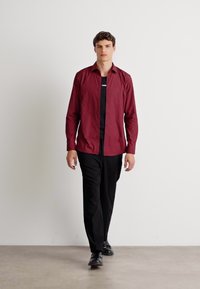 Maronfarvet skjorte med knapper, sort tætsiddende t-shirt under, sorte tapered bukser og sorte sko. Glat stof med en struktureret pasform.