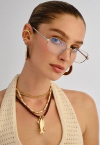 Γυναίκα με μαλλιά χτενισμένα προς τα πίσω, που φοράει ορθογώνια γυαλιά, χρυσούς και ξύλινους κρίκους σκουλαρίκια και πολυστρωματικά κολιέ, συμπεριλαμβανομένου ενός χρυσού μενταγιόν σε σχήμα ψαριού.