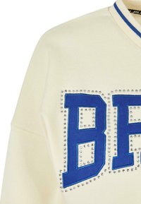 Cream-colored sweatshirt with blue embroidered letters "BF" and silver studs outlining the design. Ribbed blue collar. Soft texture.