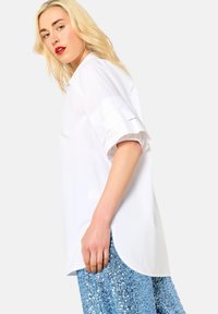Camicia bianca oversize con maniche corte arricciate. Tessuto liscio con orlo curvo. Indossata con una gonna blu scintillante ricoperta di paillettes.