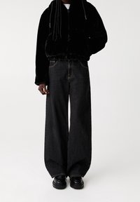 Persona vestida con pantalones vaqueros anchos negros, zapatos gruesos negros y una chaqueta de cremallera negra de felpa frente a un fondo neutro.