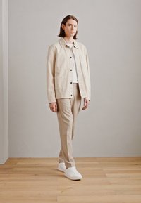 Veste en coton beige avec fermeture à boutons, deux poches avant, associée à un pantalon beige clair et des baskets blanches, sur un fond neutre.