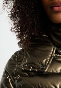 Chaqueta acolchada de color verde oliva y brillante con un cuello alto. Cuenta con botones a presión y textura acolchada. Cabello rizado visible en el fondo.