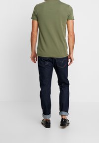 T-shirt a maniche corte verde oliva abbinato a jeans slim-fit blu scuro. I jeans hanno le estremità risvolte e una cucitura sottile sui taschini posteriori. Scarpe nere.