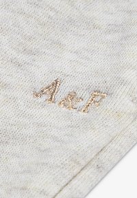 Svetlosivá textúrovaná tkanina má vyšívané iniciály "A&F" zlatou niťou, umiestnené v strede materiálu, čo vyjadruje neformálny dizajn.