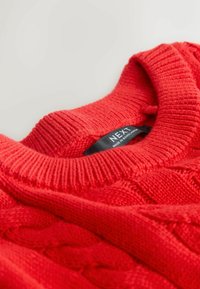 Punane õmmeldud kampsun ribidega ümarate kaelustega ja nööride kudumise mustriga; omab musta sildi, millel on tekst "NEXT" ja hooldusjuhised.