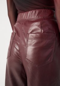 Bordeaux leren broek met een hoge taille en twee achterzakken met gestikt detail, die een gladde, glanzende textuur toont.