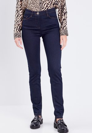 Jean skinny bleu foncé taille haute, avec des coutures contrastées et un bouton métallique à l'avant. Associé à une chemise à motifs et des chaussures brillantes.