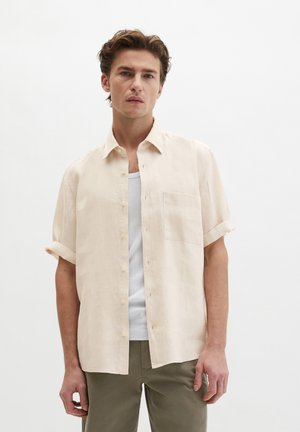 Młody mężczyzna w beżowej koszuli z krótkim rękawem zapinanej na guziki, nałożonej na biały tank top oraz oliwkowo-zielone spodnie, stojący na tle jednolitego białego tła.