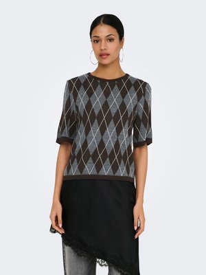 Žena oblečená v hnědém a šedém krátkém svetru s kosočtverečným vzorem a černé asymetrické sukni vrstvené přes džíny, stojící na bílém pozadí.