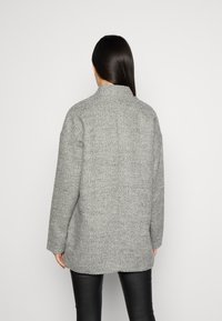 Grauer Mantel aus Wollmischgewebe mit Fischgrätmuster, hohem Kragen, lockerem Schnitt und abgesenkten Schultern. Strukturierte Oberfläche und lange Ärmel.