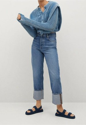Person iført en struktureret blå strikket sweater, højtaljede optrukne jeans og navyblå sandaler, stående mod en ensfarvet lys baggrund.