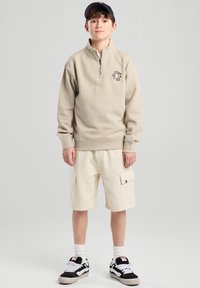 Beige zip-up sweatshirt with logo, paired with white cargo shorts and black sneakers with white accents. Model has a black cap.