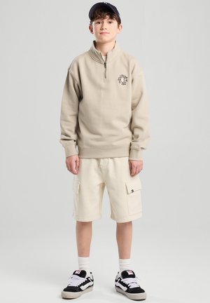 Beige zip-up sweatshirt with logo, paired with white cargo shorts and black sneakers with white accents. Model has a black cap.