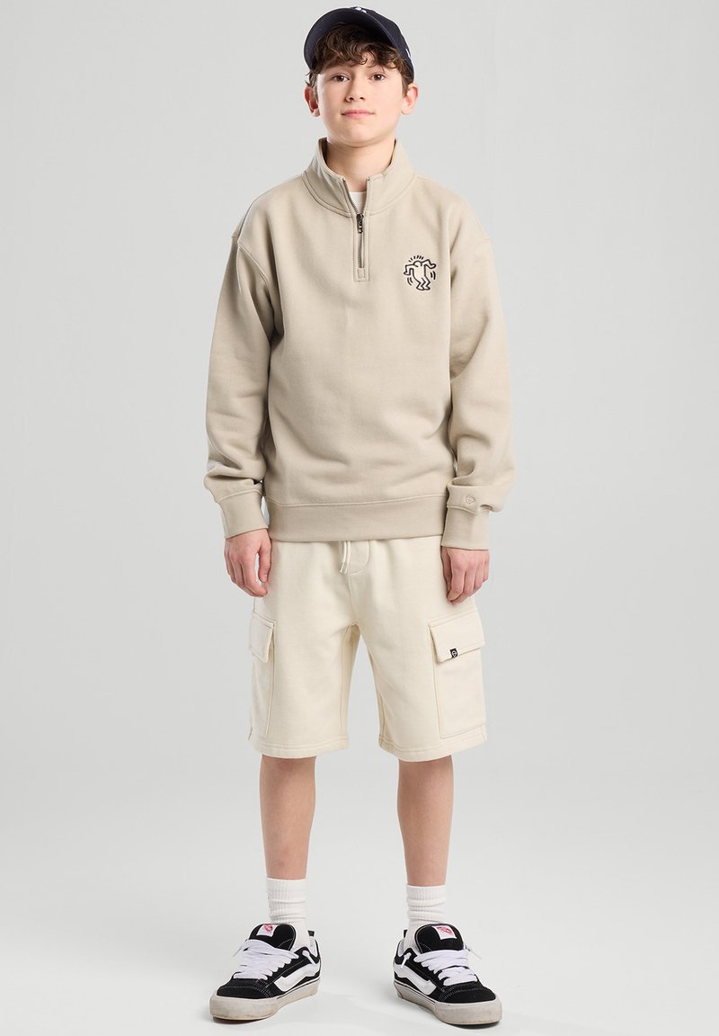 Beige zip-up sweatshirt with logo, paired with white cargo shorts and black sneakers with white accents. Model has a black cap.