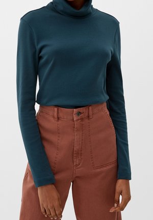 Personne portant un col roulé à manches longues couleur sarcelle rentré dans un pantalon taille haute de couleur rouille avec des poches plaquées à l'avant.