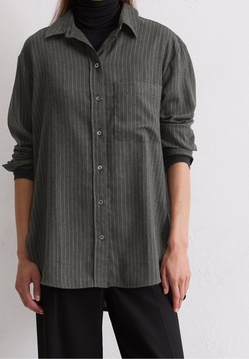 Personne portant une chemise boutonnée à fines rayures vert foncé sur un col roulé noir avec un pantalon noir, debout devant un mur blanc uni.