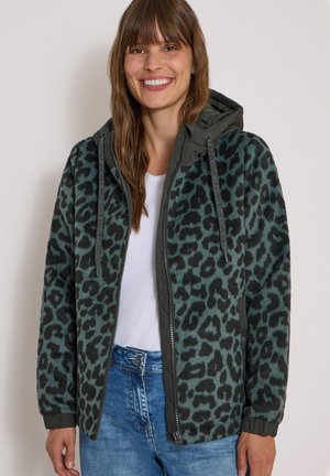 Kvinde smilende iført en grøn og sort leopardprint lynlåshættejakke over en hvid skjorte og blå jeans mod en ensfarvet baggrund.