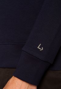 Granatowy materiał bluzy z ribbowymi mankietami. Posiada jasnoszare logo na rękawie. Tekstura jest gładka i miękka, o luźnym designie.