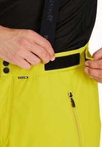 Ljust gula skidbyxor med en slät yta, justerbart midjeband med kardborreknäppning och ficka med dragkedja med svarta detaljer.