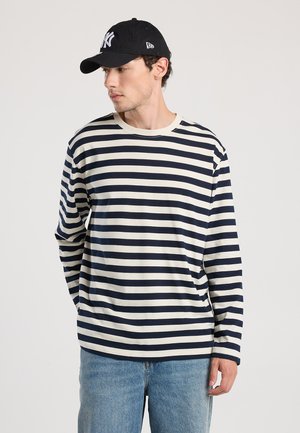 Long-sleeve striped shirt in navy and cream cotton, featuring a crew neck. Worn with a black cap. Casual fit and simple pattern.