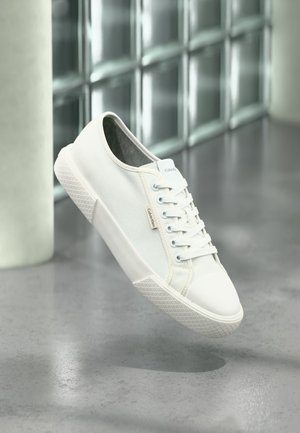Λευκό χαμηλό sneaker Calvin Klein με κορδόνια, αιωρούμενο πάνω από γκρίζο γυαλισμένο πάτωμα και φόντο από τοιχοποιία με γυάλινους κύβους.