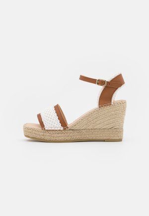 Sandale compensée marron et blanche avec bride de cheville, semelle plateforme en corde de jute et bride avant texturée, présentée sur fond blanc uni.