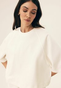 Weißes, kurzärmeliges Sweatshirt mit lässiger Passform, rundem Halsausschnitt und sichtbaren Nähten. Weicher Stoff mit glatter Textur.