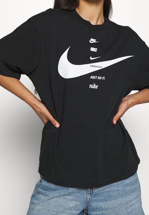 Osoba nosząca czarną koszulkę Nike z kilkoma białymi logo typu swoosh oraz napisami „SWOOSH,” „JUST DO IT.,” i „nike” umieszczonymi na środku z przodu.