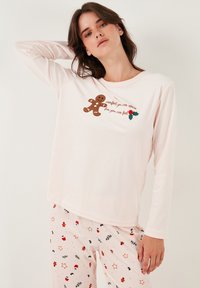 Vaaleanpunainen pitkähihainen pyjamapaita, jossa on piparkakkumiehen grafiikka ja teksti. Yhteensopivissa pyjamahousuissa on piparkakku- ja juhlapainatus.