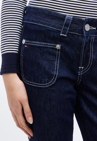 Χέρι που ακουμπά δίπλα στην τσέπη από σκούρο μπλε τζιν με λευκά ράμματα, φορεμένο με μπλουζάκι μακρυμάνικο σε ναυτικό μπλε και λευκές ρίγες.