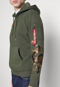 Sudadera verde oliva con un bolsillo frontal, que presenta un gráfico de dragón en la manga izquierda y un tira de cremallera roja con texto. Fabricada en tejido suave.