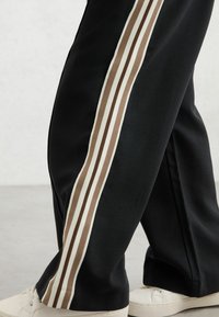Pantalon de survêtement noir avec trois bandes verticales crème et marron sur les côtés, en matière douce, associé à des baskets blanches.