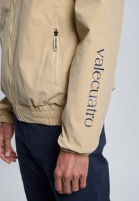 Chaqueta bomber beige con puños elásticos, bolsillo lateral con cremallera y logo "vale-cuatro" en azul marino en la manga. Textura suave y diseño casual.