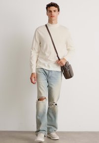 Maglione lavorato a maglia crema, jeans strappati azzurri chiari e sneakers bianche. Borsa a tracolla marrone con tracolla regolabile. Outfit casual.