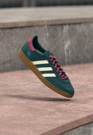 Πράσινο αθλητικό παπούτσι Adidas με λευκές ρίγες, μπορντό κορδόνια και λαστιχένια σόλα που αιωρείται πάνω από μια πέτρινη επιφάνεια σε εξωτερικό χώρο.