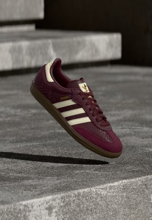 Maroon Adidas Samba sneaker met crèmekleurige strepen en gumzool, hangend boven betonnen treden in zacht licht.
