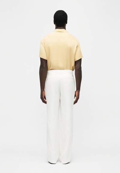 Homme portant une chemise à manches courtes jaune clair rentrée dans un pantalon blanc taille haute à jambes larges, debout de dos sur un fond uni.