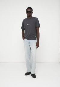 Pelēks kokvilnas t-krekls ar īsām piedurknēm un vieglu logo, apvienots ar gaiši ziliem taisnām kājām džinsiem un melnām kedām, valkājot saulesbrilles.